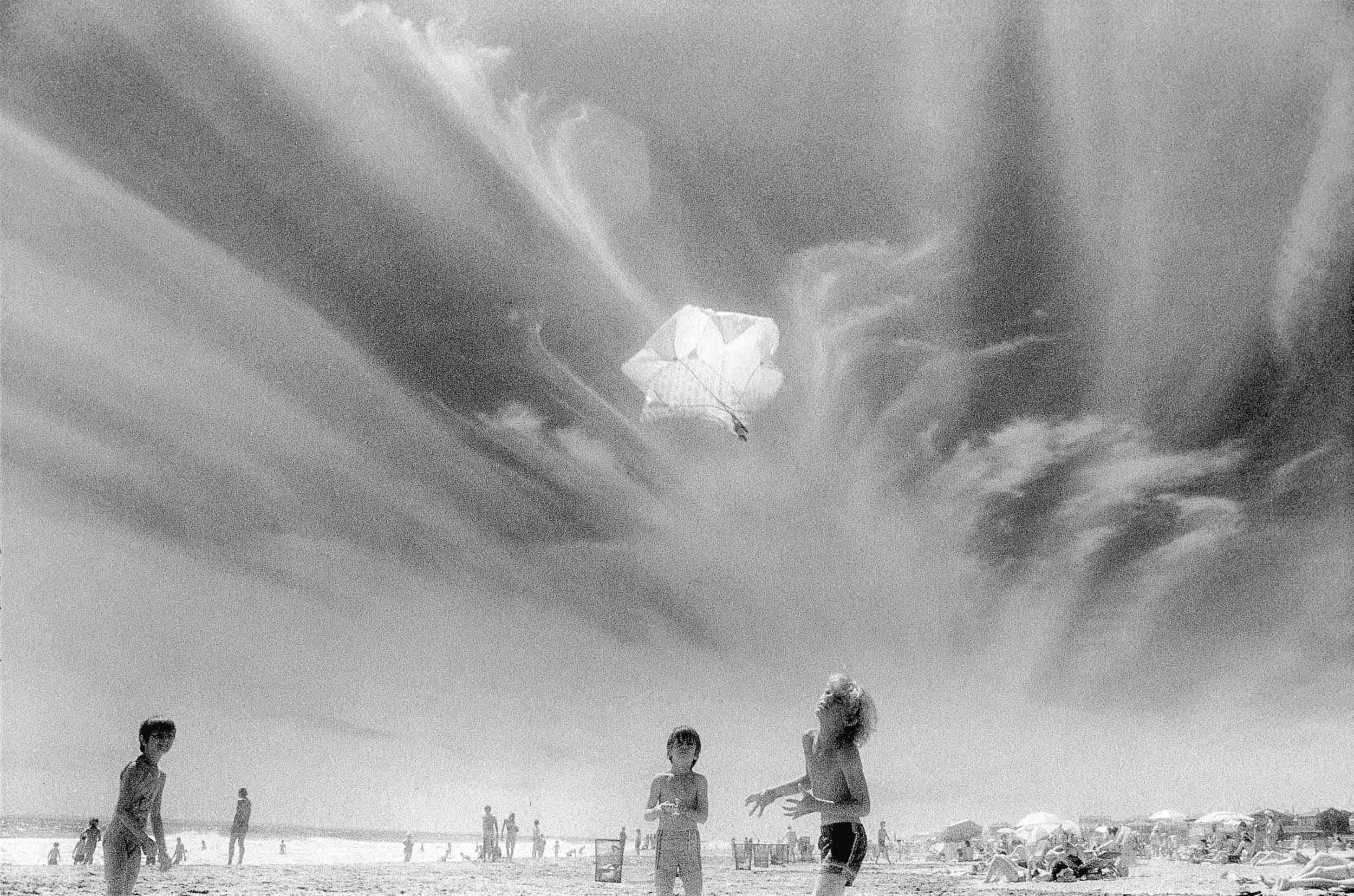 Barbara Alper- Photo of kids tossing up a toy parachute into clouds at Rockaway Beach NYC