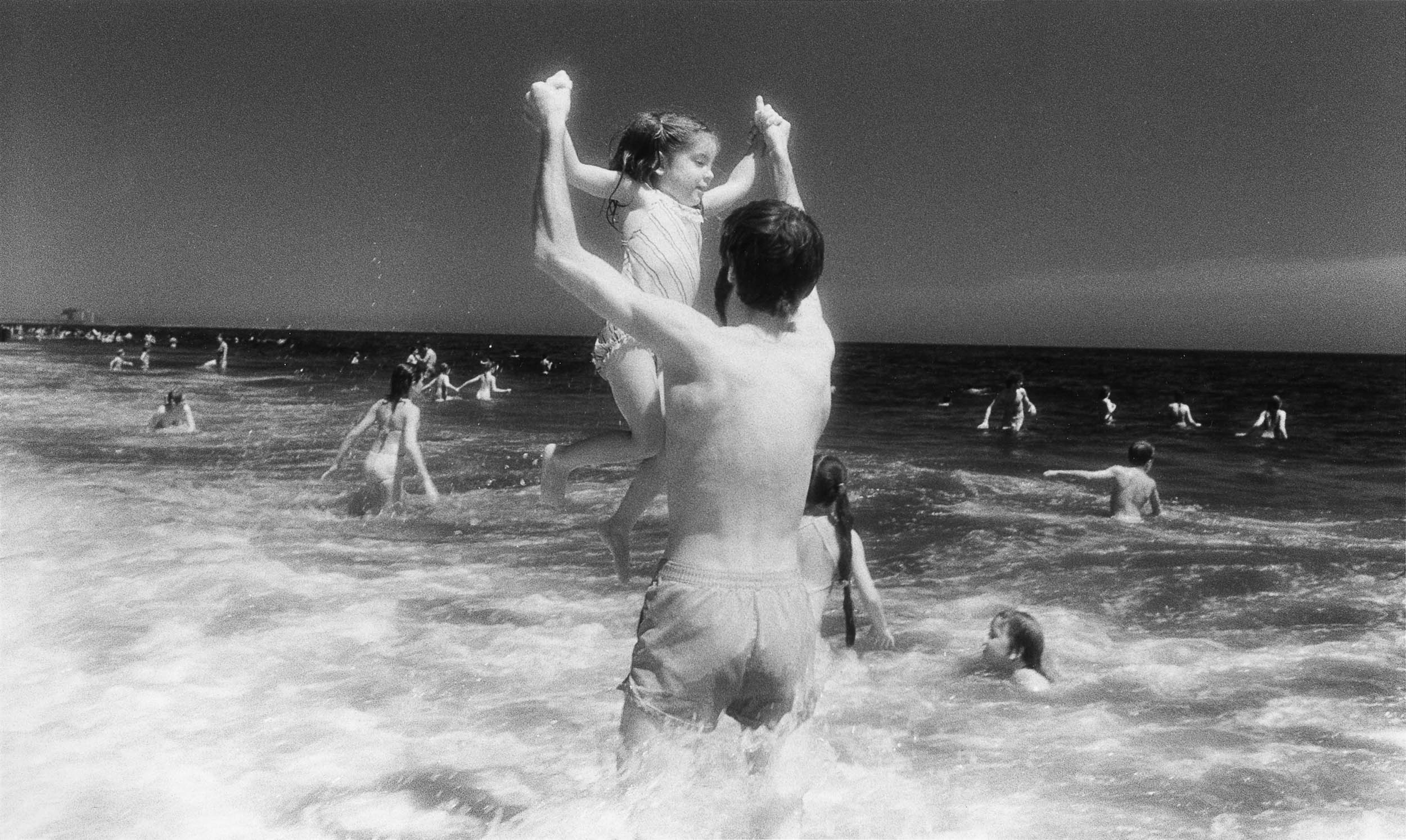 Barbara Alper- Photo of man holding girl up in the air in the water at Rockaway Beach NYC
